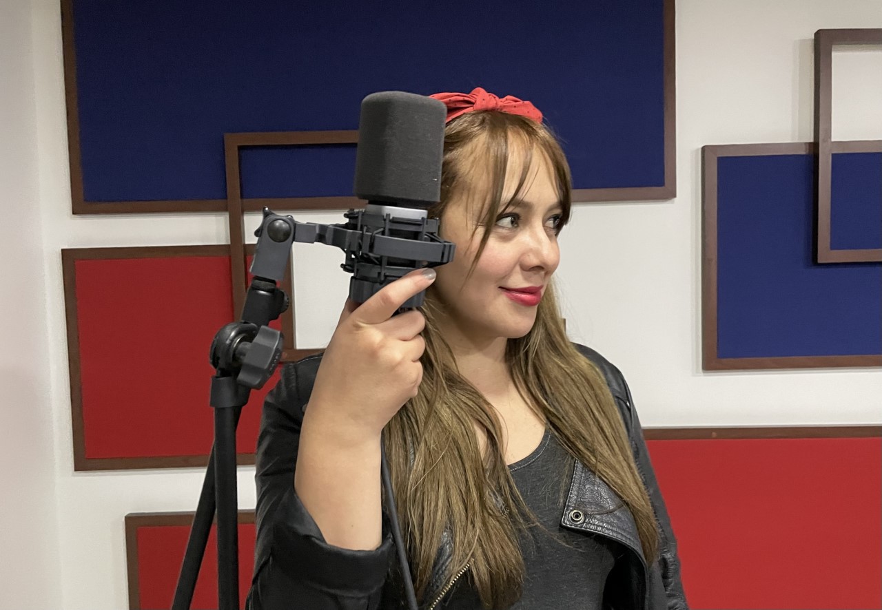 Adriana sostiene el micrófono con su mano derecha, ella mira hacia su lado izquierdo
