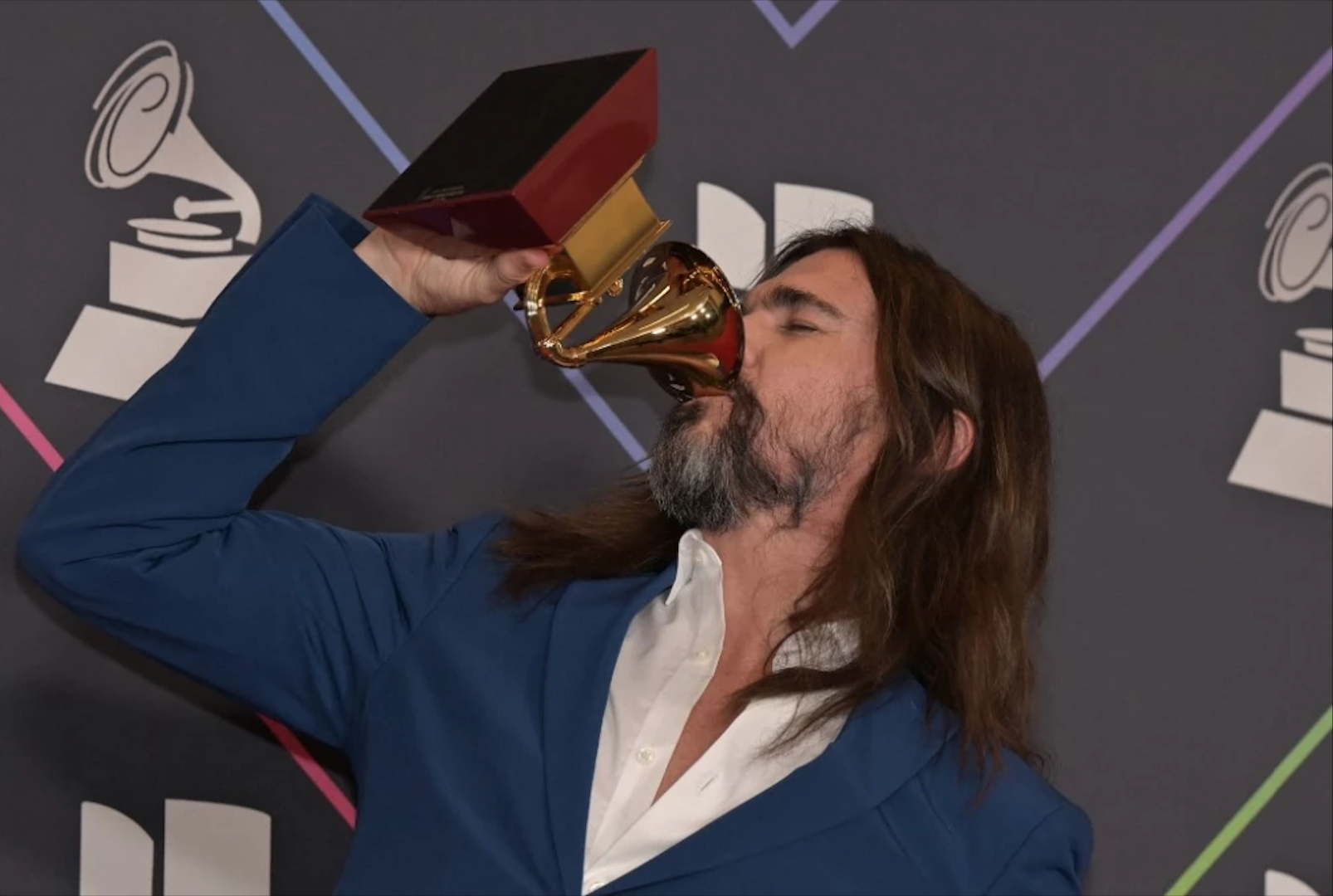 Juanes viste un traje azul y camisa blanca, en su mano sostiene un Grammy ganado por el álbum orígenes, él simula que es una copa y que está bebiendo algo de ella