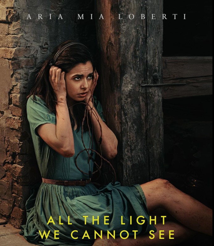 Aria Mia Loberti en el papel de Marie-Laure Leblanc, ella viste un vestido color verde,  está en el suelo, tiene varias raspaduras en sus piernas y brazos, ella sostiene con sus manos unos audifonos que cubren su orejas 