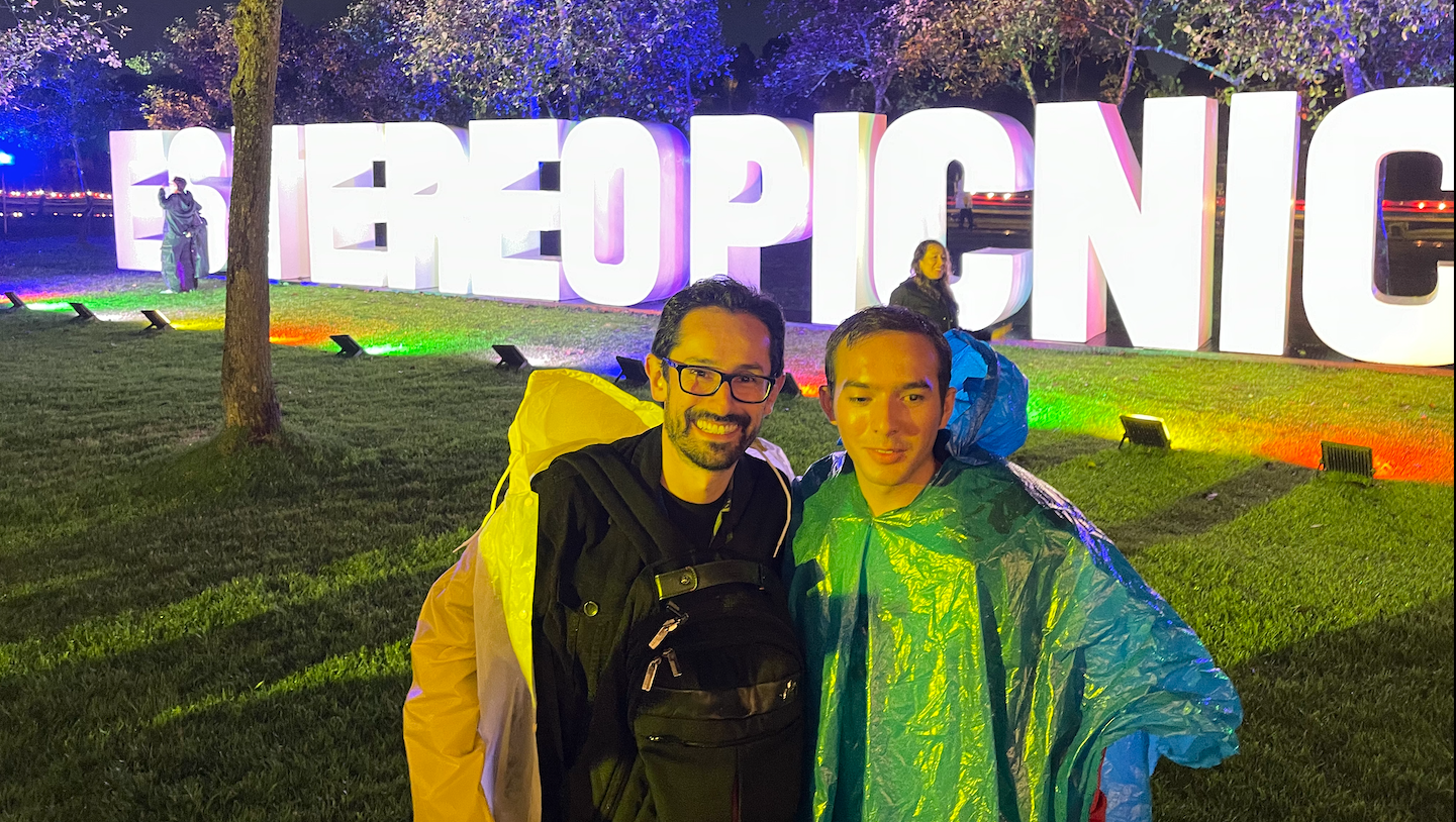 camilo garnica  junto a Henry Díaz usan impermeables atrás en letras blancas hay un letrero que dice Estéreo Picnic la foto de realizó de noche 