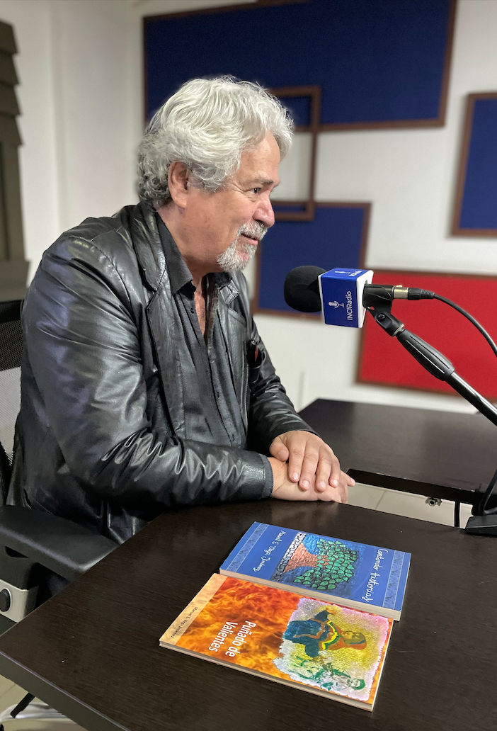 manuel está de medio lado frente al micrófono a su derecha están dos de sus libros, uno de color naranja se llama puñado de valientes y el otro de color azul se llama contando historias