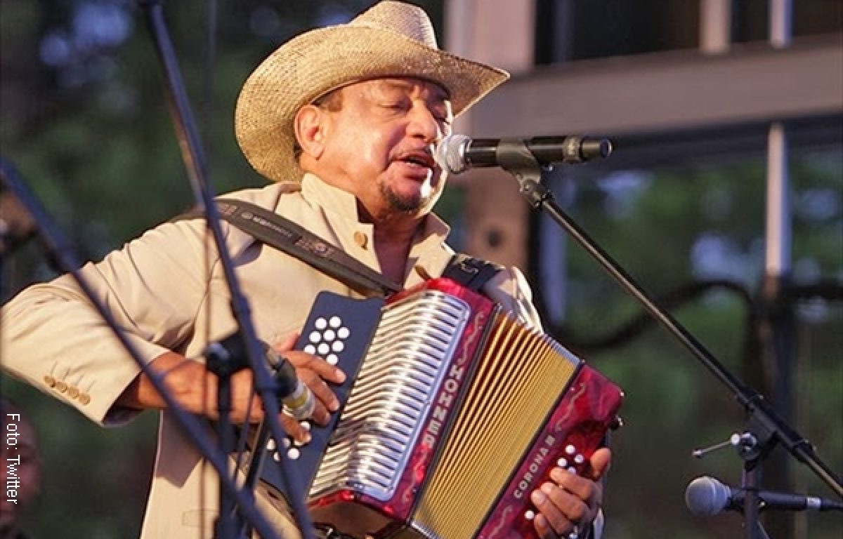 Lizandro viste de blanco con un sombrero tipico del mismo color en sus manos sostiene un acordeon 