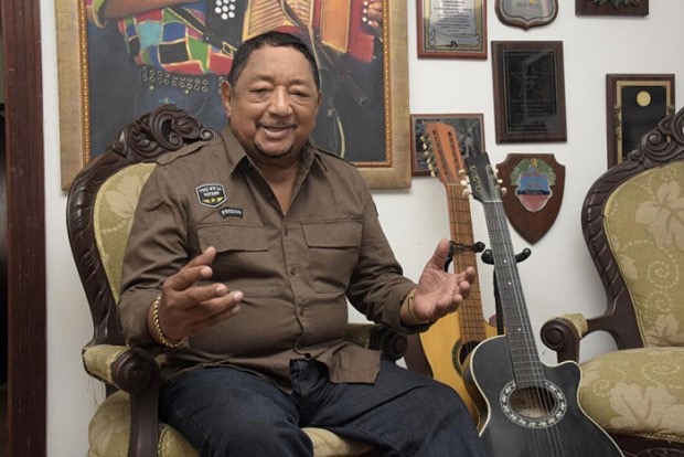 Lisandro está en la sala viste una camisa de color café a su lado está un cuadro de él sosteniendo  un acordeon y dos guitarras una de seis y otra de doce cuerdas