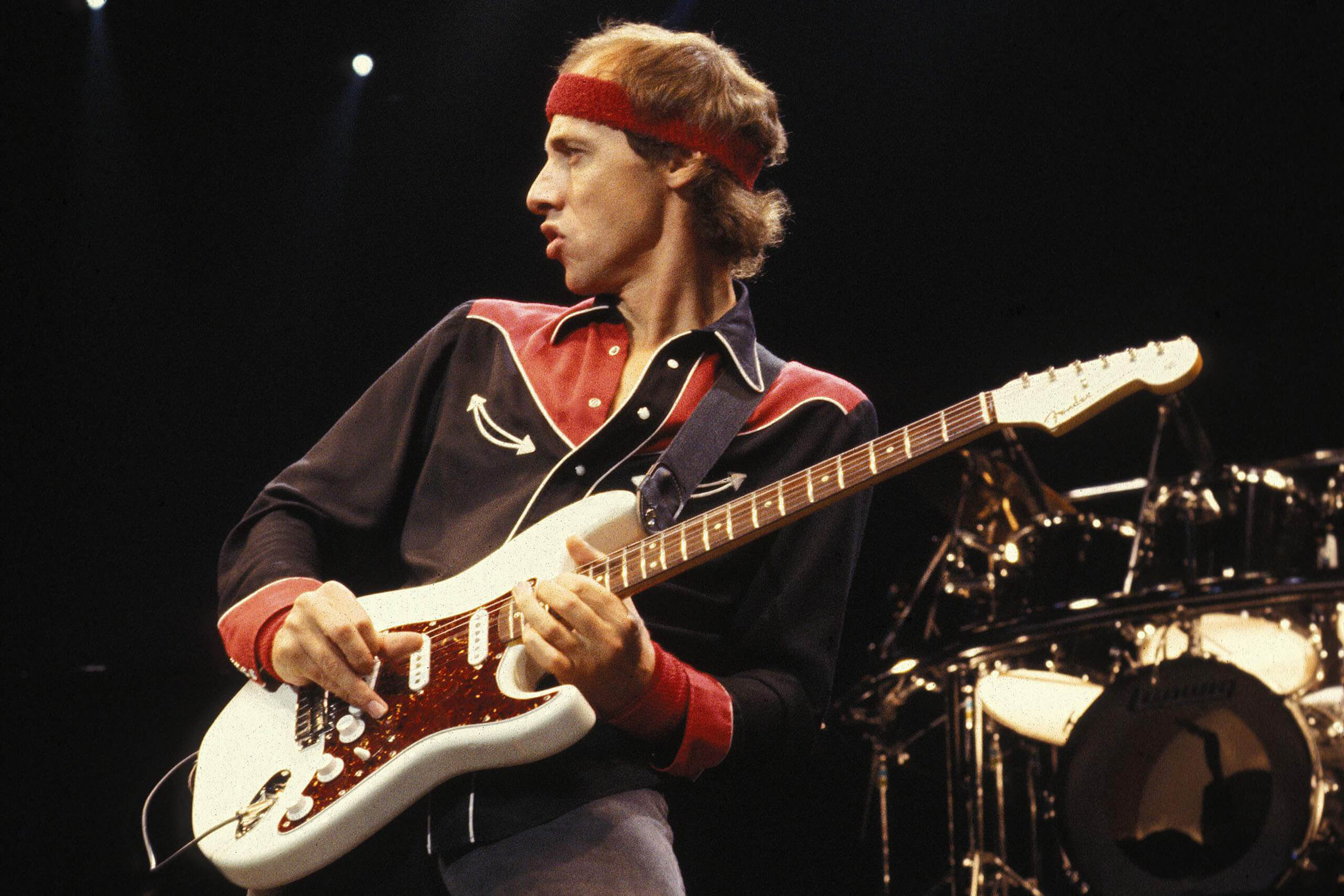 mark knopfler tocando la guitarra usa una balaca deportiva roja, usa una camisa negra y una guitarra blanca con rojo