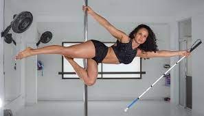 Andrea en horizontal cuelga del pole, su pierna derecha está enlongada totalmente, mientras su pierna izquierda reposa sobre la derecha, en su mano izquierda sostiene su bastón blanco  