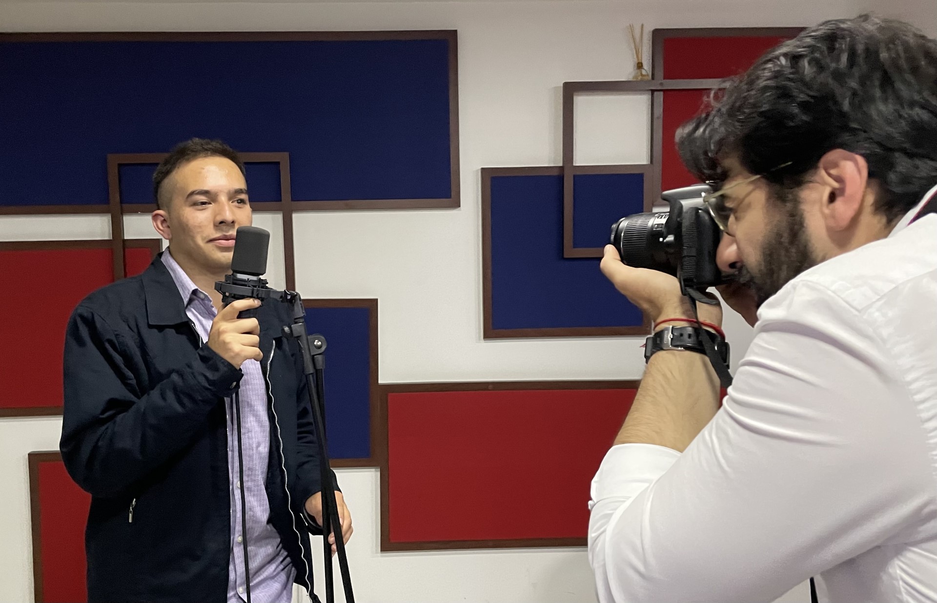 en la cabina de radio, Juan esteban sostienee una cámara de video está grabando a camilo garnica quién aparece frente al micrófono