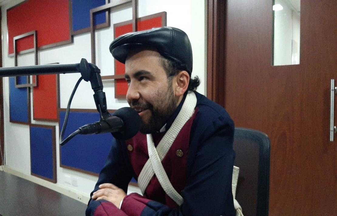 Imagen, Carlos Quintero sonríe frente al micrófono, él usa una boina de color negro, usa un abrigo de color azul y rojo,