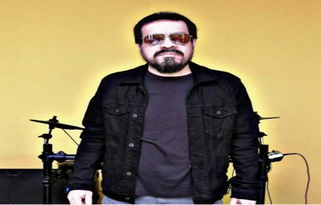 Imagen del Doctor Rock, el está frente a una batería, él viste un buzo color vinotinto y una chamarra negra, usa unos lentes de sol