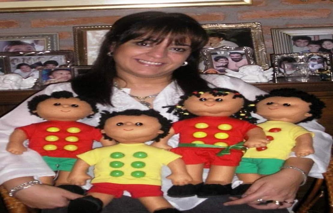 Virginia Perez sostiene en sus brazos a seis braillines, dos vestidos con camisa roja y botones que forman el signo generador de color amarillo, y dos braillines con camisa amarilla y botones de color verdes y rojos, ella sonríe