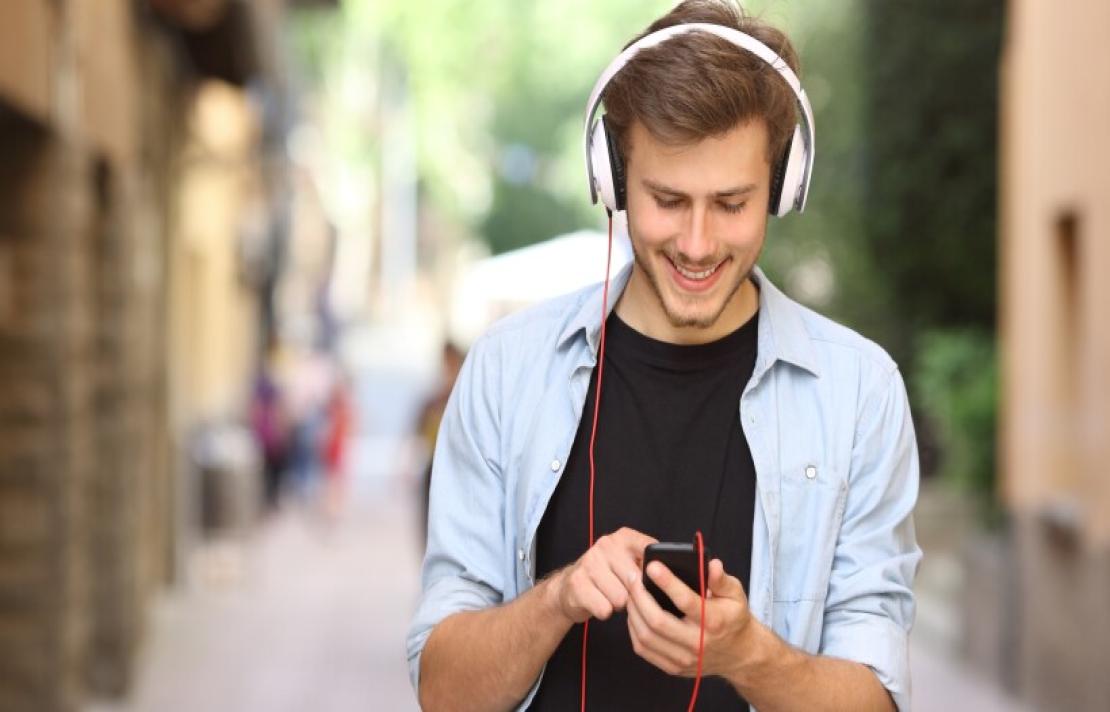 hombre joven viste una camisa azul abierta se ve una camiseta de color negro, él sostieen en sus manos un celular, mientras escucha algo con sus audifonos, él sonríe