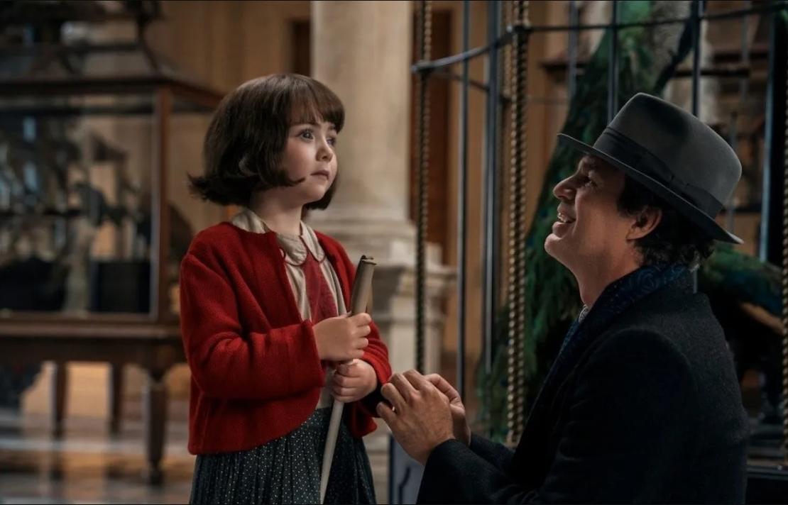 la niña Nell Sutton interpretando su papel de Marie-Laure Leblanc, sostiene su bastón mientras a su derecha está un sonriente Mark Ruffalo en su personificación de Daniel LeBlanc él está arodillado para quedar a la altura de su acompañante