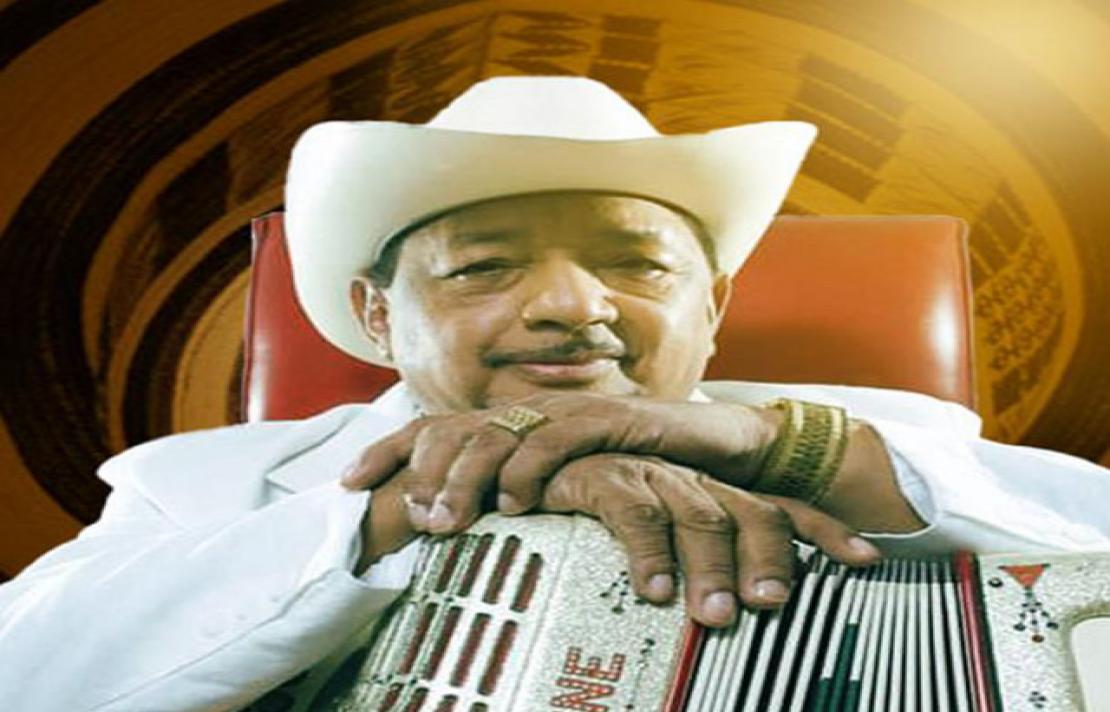 frente a un fondo animado de un sombrero vueltiao y vistiendo un traje y sombrero blanco está lisandro mesa quien posa sus manos sobre un acordeón