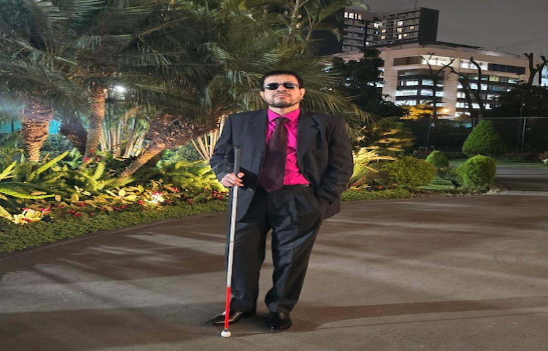 de noche, Braulio Mogollón viste un traje negro, su corbata es del mismo color, usa además una camisa de color rosado, Braulio está junto a su bastón guía,