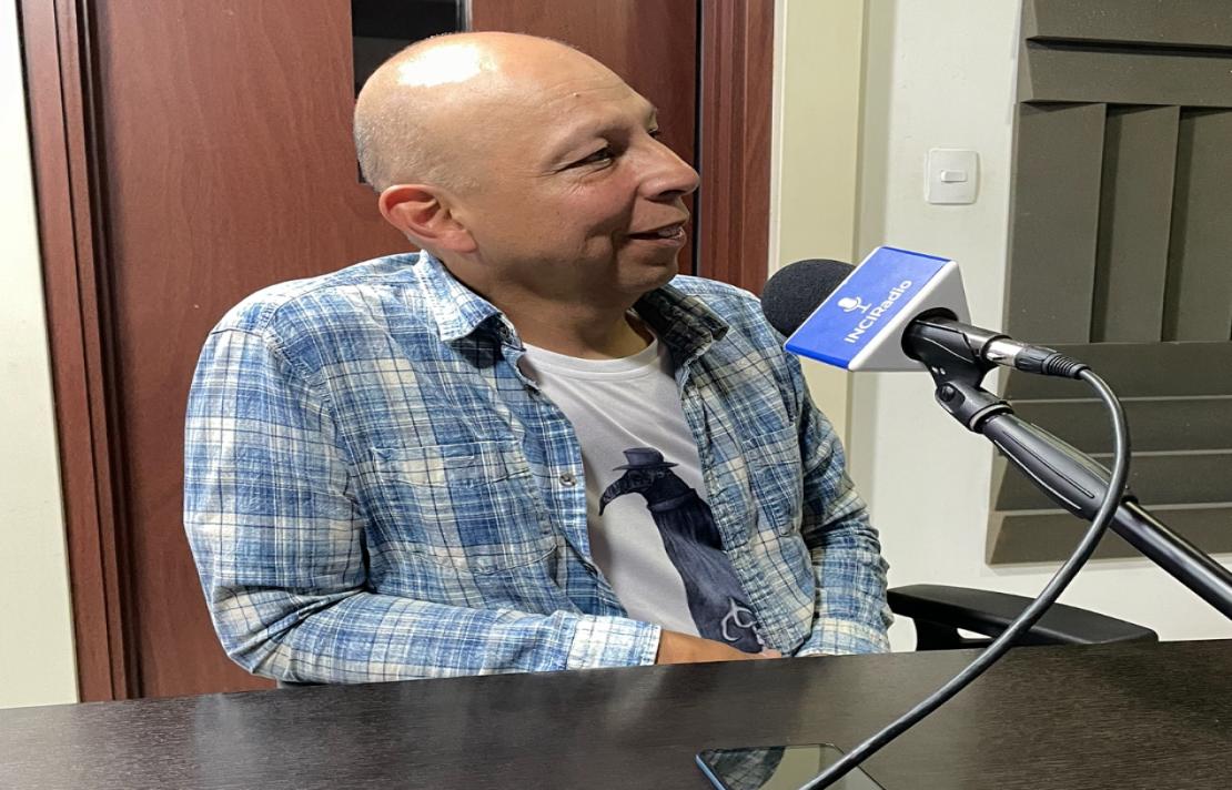 Jhon Jairo Jiménez "Jota" está frente al micrófono, el viste una camisa a cuadros y una camiseta blanca