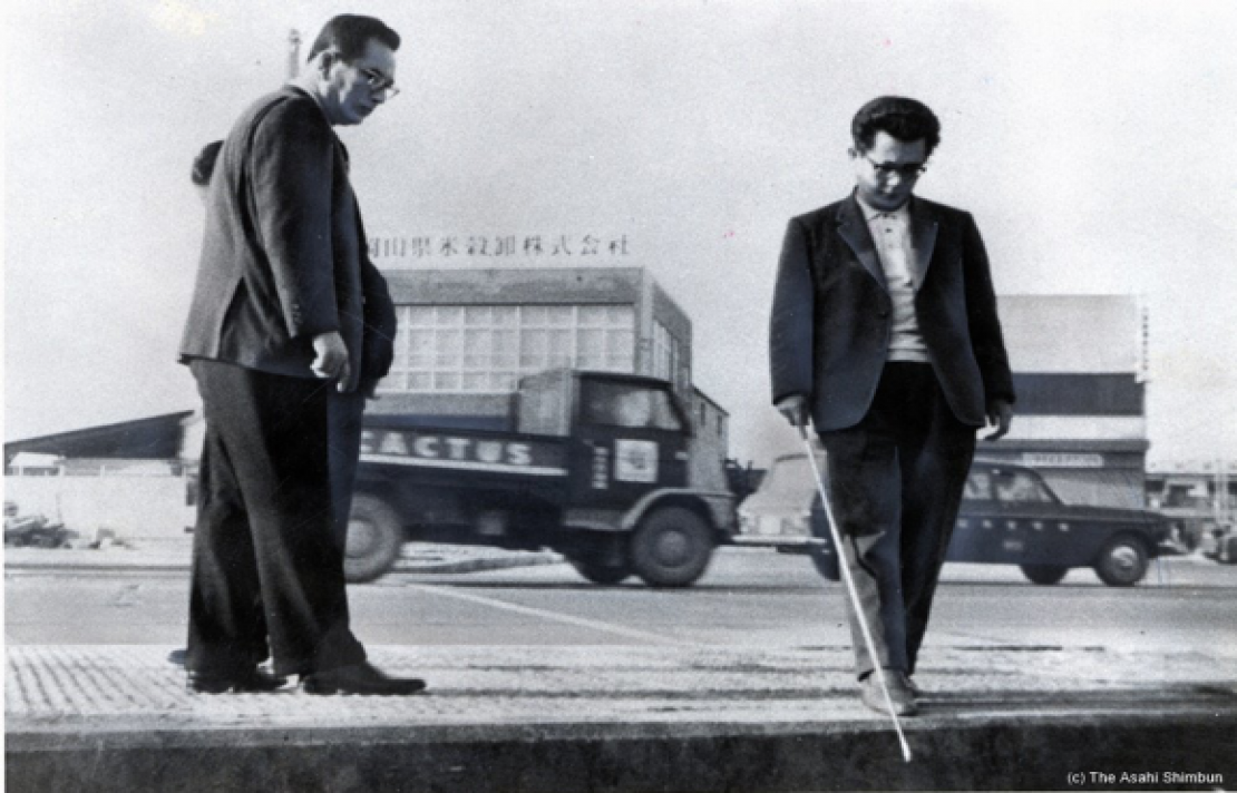 imagen antigua en Japón donde encontramos una persona caminando con su bastón blanco, una persona lo mira, de fondo se ven unos camiones y unos carros estacionados