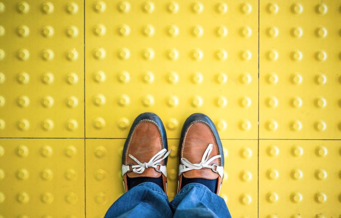 imagen de una señal podotáctile, sobre ella aparecen las piernas de una persona que viste un jean de color azul, sus zapatos son café